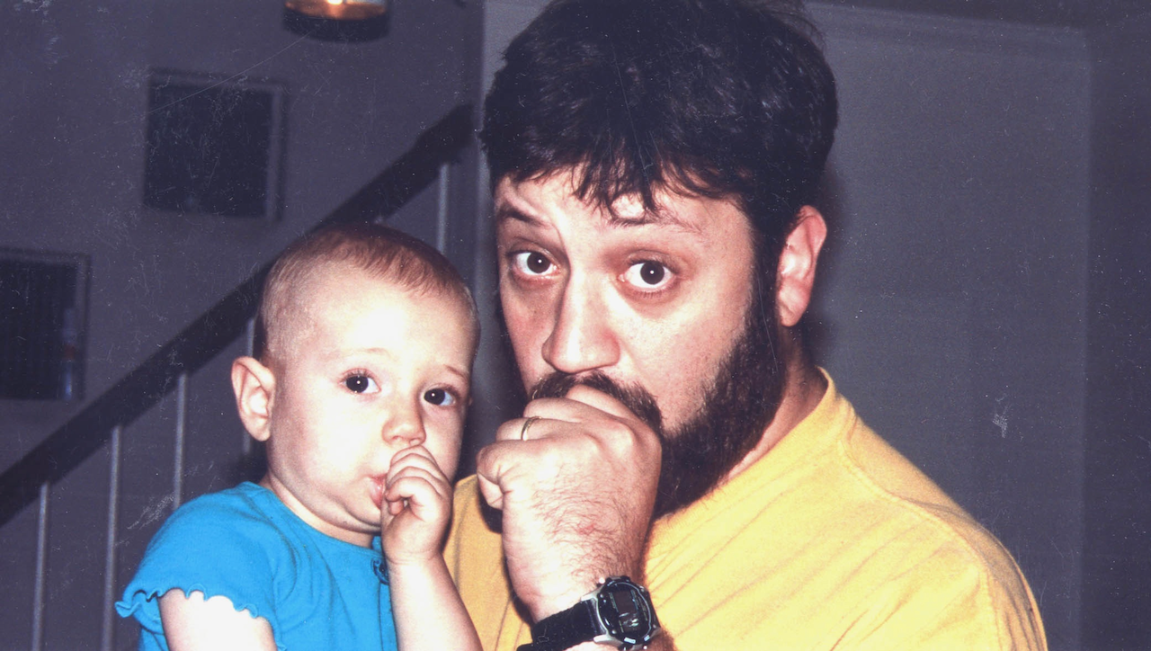 Bearded father in a yellow shirt holding a baby indoors near a staircase